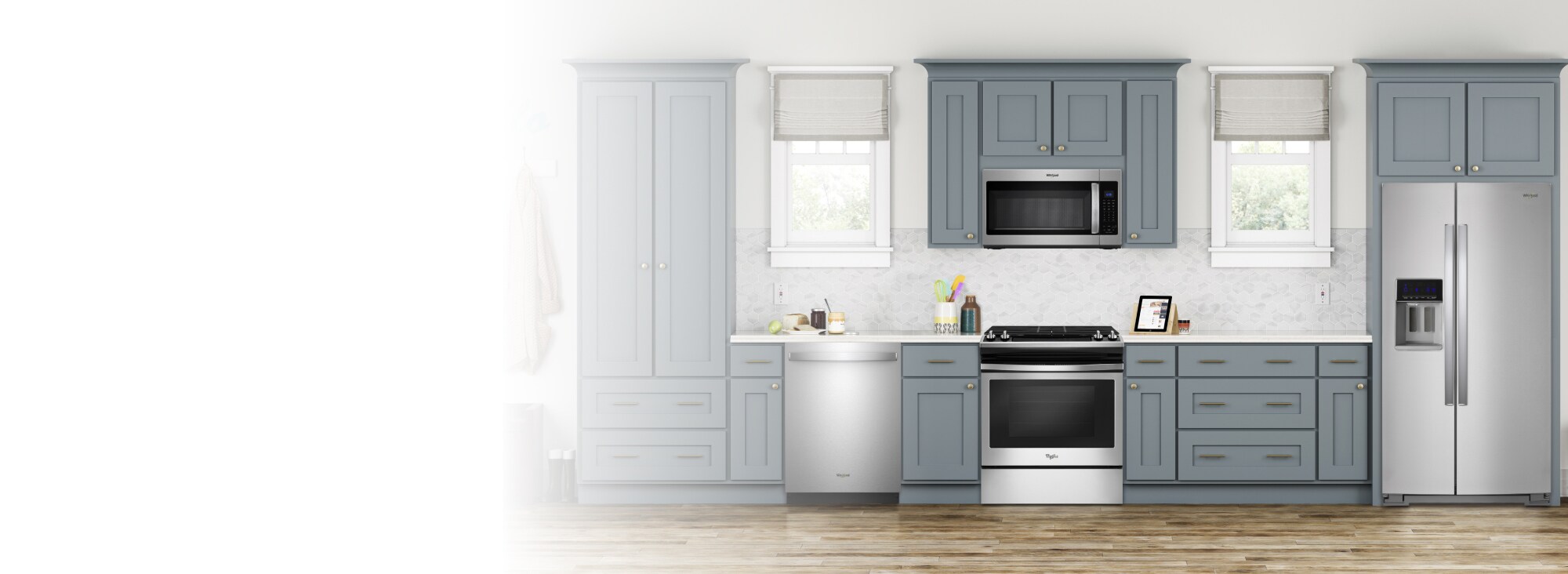 A kitchen with grey cabinets and two windows. There are several appliances, including a Whirlpool Range, Whirlpool Over-the-Range Microwave, Whirlpool Dishwasher and Whirlpool Refrigerator