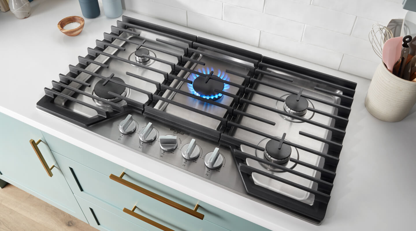 Gas cooktop above light blue cabinetry with fifth middle burner lit