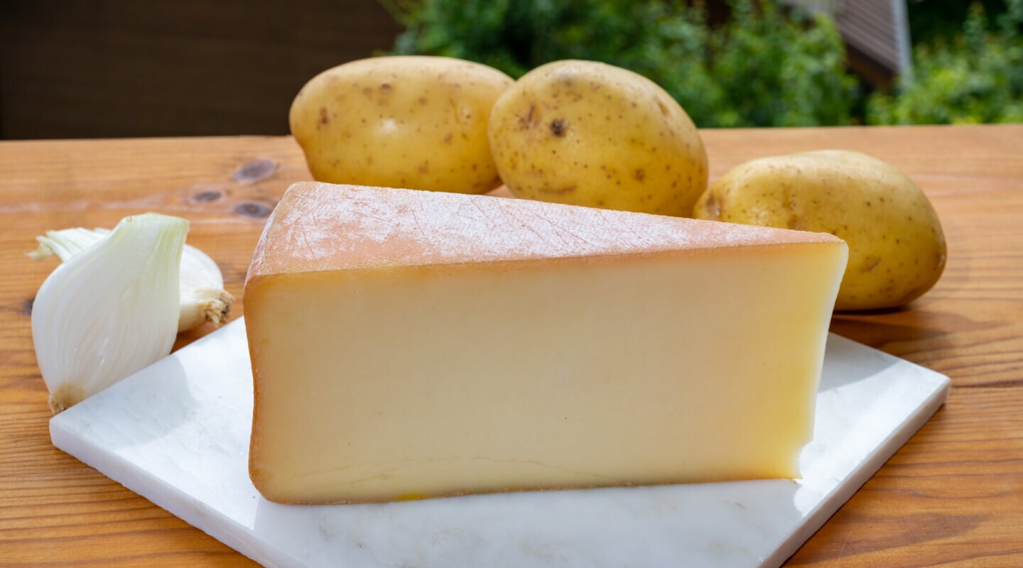 A wedge of cheese on a cutting board with three potatoes behind it
