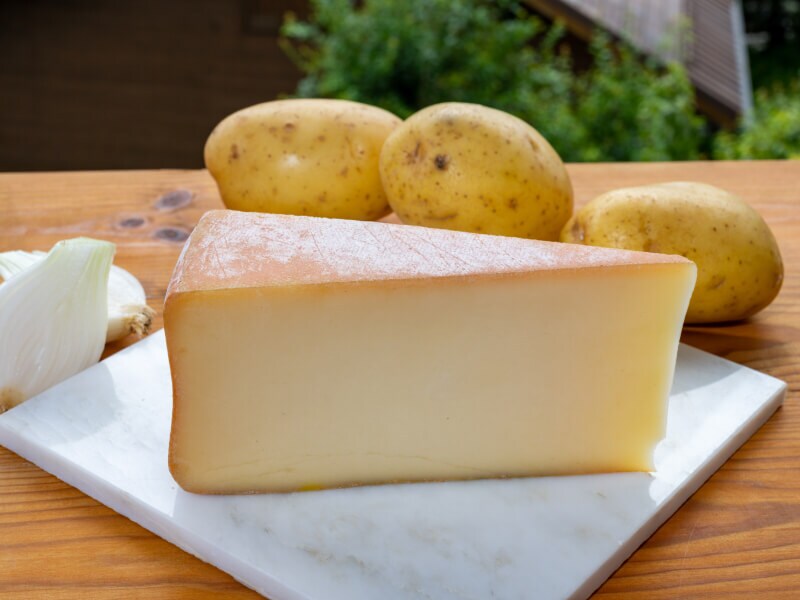 A wedge of cheese on a cutting board with three potatoes behind it