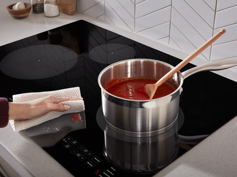 Person wiping cooktop next to burner with saucepan Person wiping cooktop next to burner with saucepan