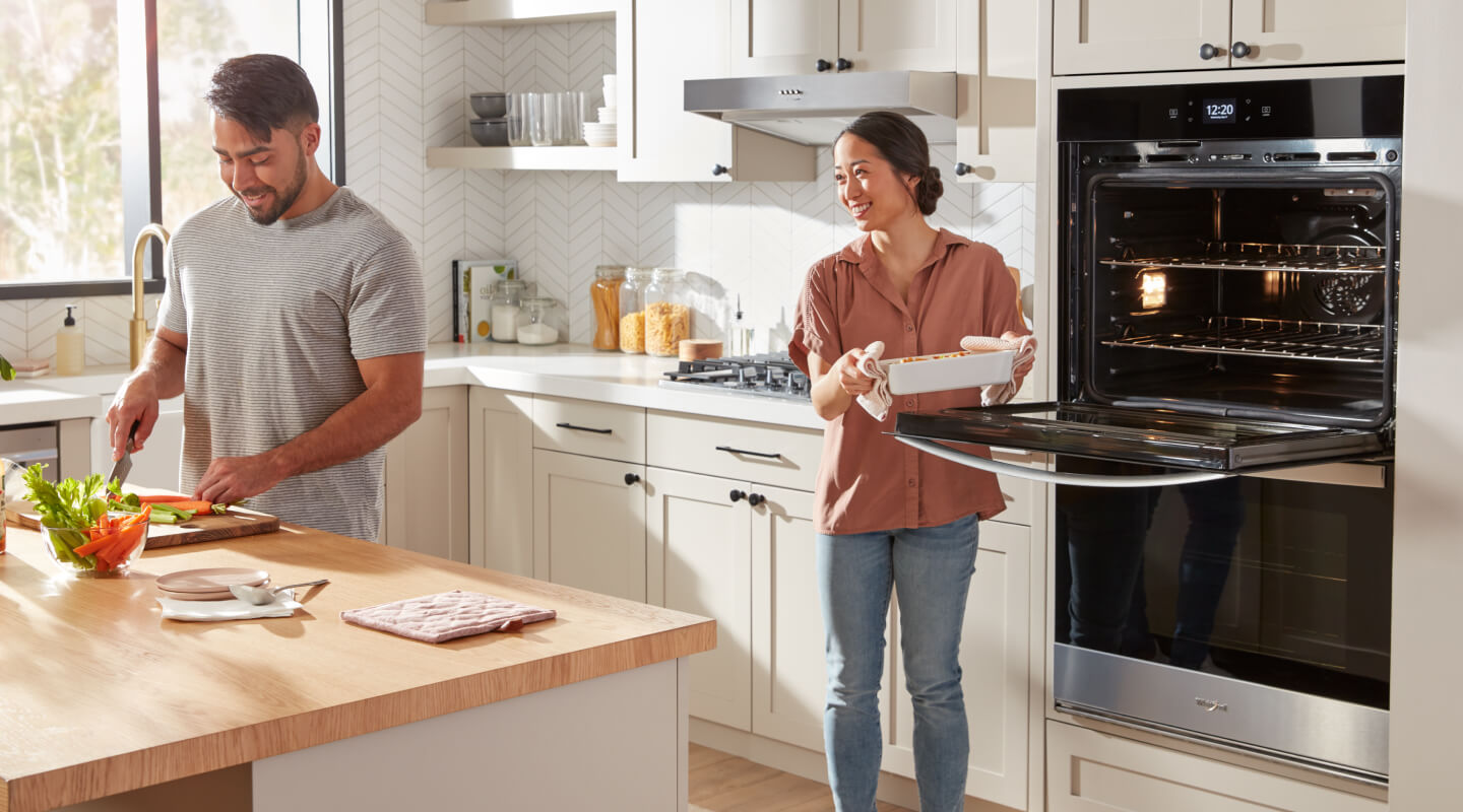 A man and woman cooking in their kitchen and pulling food out of their double wall oven