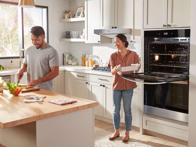 A man and woman cooking in their kitchen and pulling food out of their double wall oven