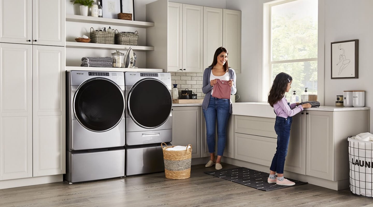 People folding clothes next to a Whirlpool® Washer and Dryer set in a modern laundry room People folding clothes next to a Whirlpool® Washer and Dryer set in a modern laundry room
