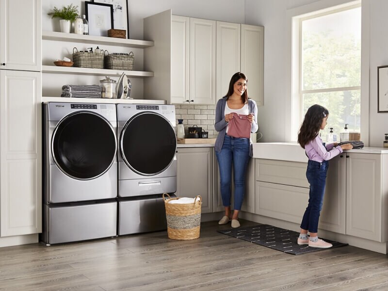 People folding clothes next to a Whirlpool® Washer and Dryer set in a modern laundry room People folding clothes next to a Whirlpool® Washer and Dryer set in a modern laundry room