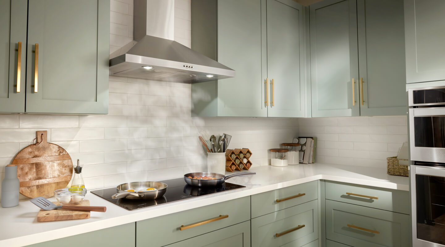Cooktop with steam rising towards the range hood in a kitchen with light green cabinets and gold handles