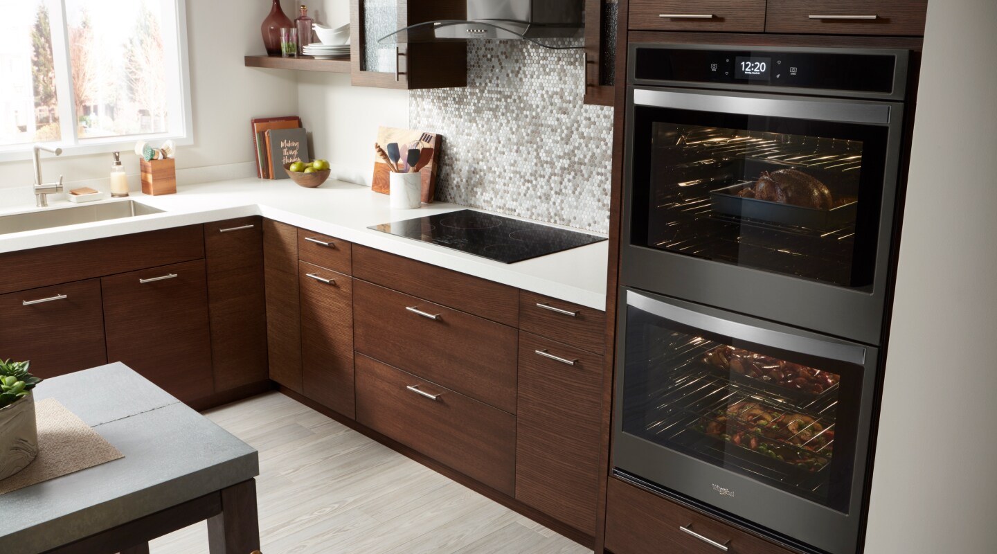 Double wall oven and an electric flat top range in a kitchen with dark wood cabinets and neutral tile backsplash