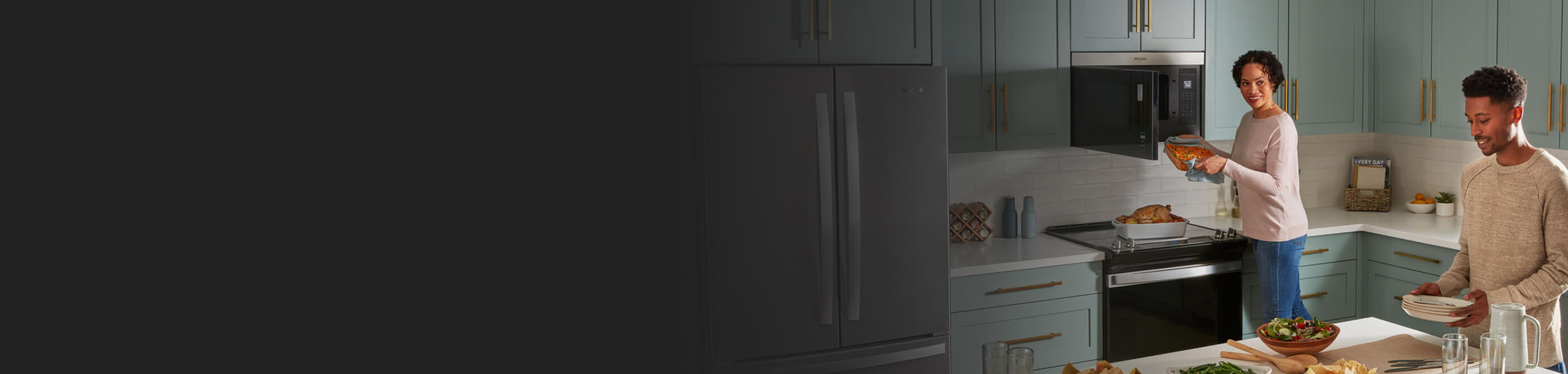 A couple making dinner in a sea green kitchen with one of them pulling a casserole dish out of a microwave