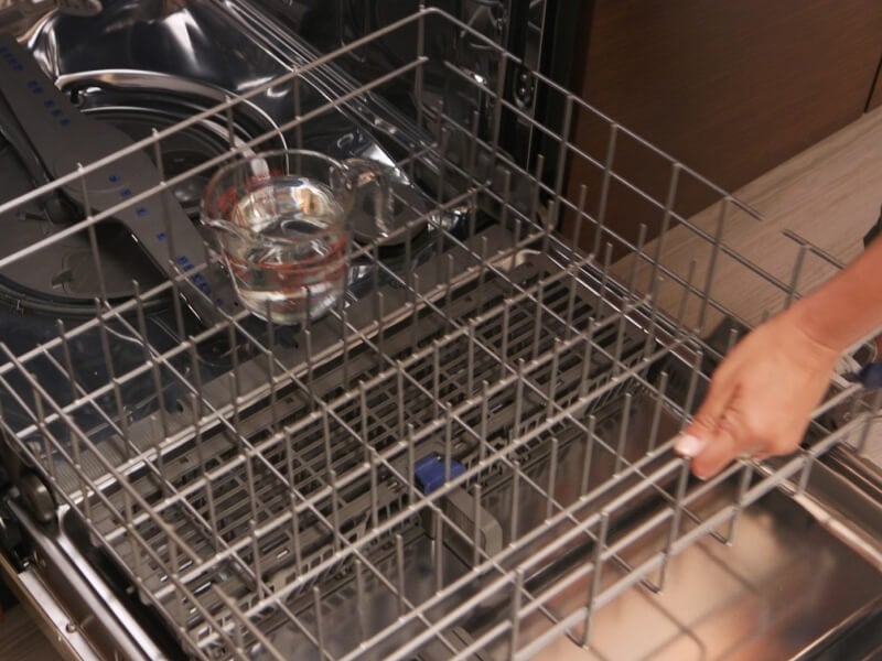 A person adding a measuring cup of vinegar to a dishwasher rack