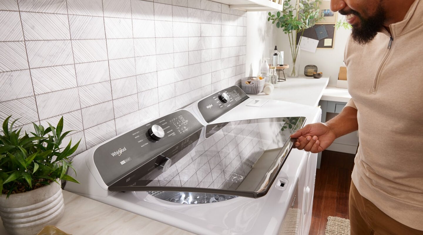 Person opening a white Whirlpool® Top Load Washer that is next to a white dryer in a laundry room Person opening a white Whirlpool® Top Load Washer that is next to a white dryer in a laundry room