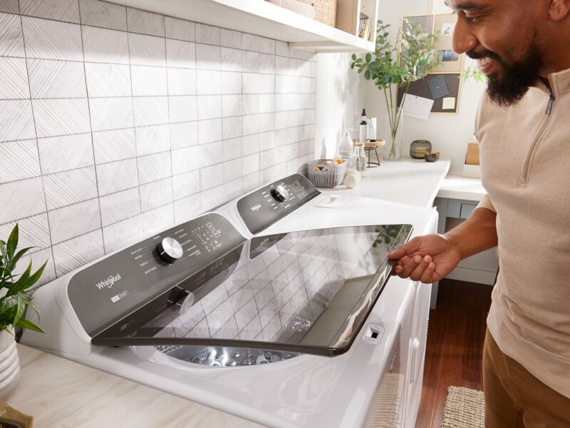 Person opening a white Whirlpool® Top Load Washer that is next to a white dryer in a laundry room Person opening a white Whirlpool® Top Load Washer that is next to a white dryer in a laundry room