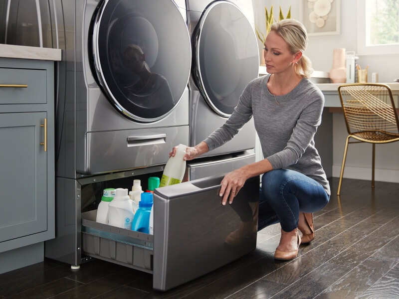 A person putting away detergent in a laundry pedestal that’s under a gray front load washer and next to a front load dryer A person putting away detergent in a laundry pedestal that’s under a gray front load washer and next to a front load dryer