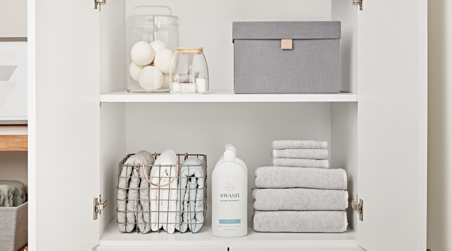 Swash® Laundry Detergent on the bottom shelf of a white cabinet between stacks of gray towels Swash® Laundry Detergent on the bottom shelf of a white cabinet between stacks of gray towels