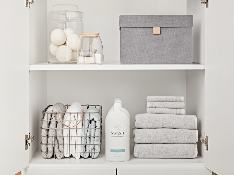 Swash® Laundry Detergent on the bottom shelf of a white cabinet between stacks of gray towels Swash® Laundry Detergent on the bottom shelf of a white cabinet between stacks of gray towels