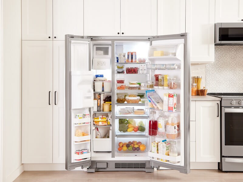 An open, side-by-side fridge filled with food and surrounded by white cabinets