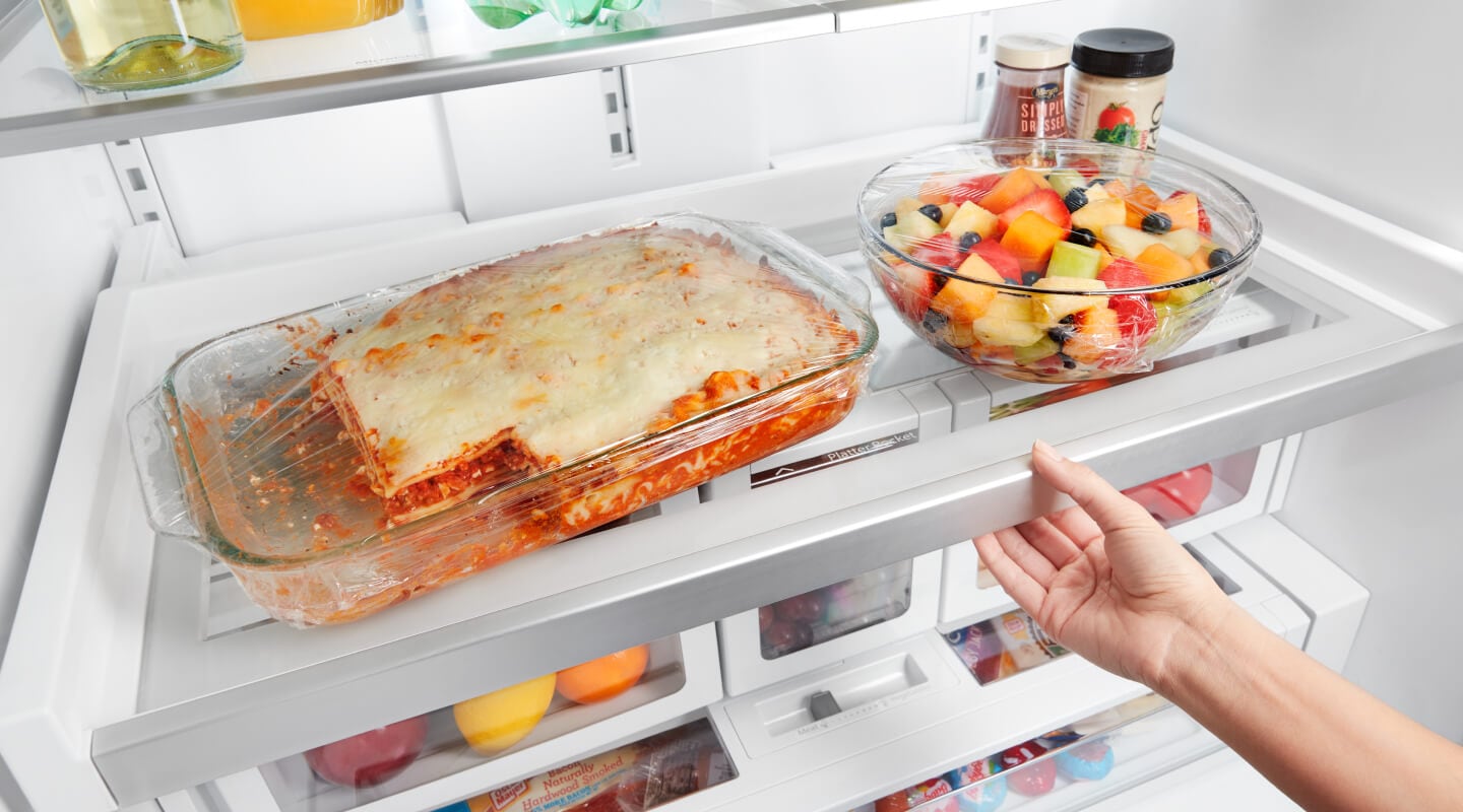 A lasagna and a fruit bowl sitting on a fridge shelf A lasagna and a fruit bowl sitting on a fridge shelf