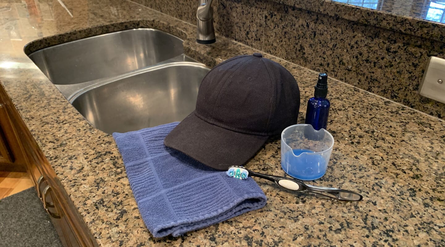 A black cloth baseball cap on a countertop with a toothbrush, towel and cleaning supplies