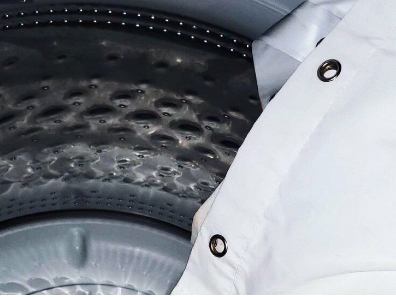 A white shower curtain being placed in a washer. A white shower curtain being placed in a washer.