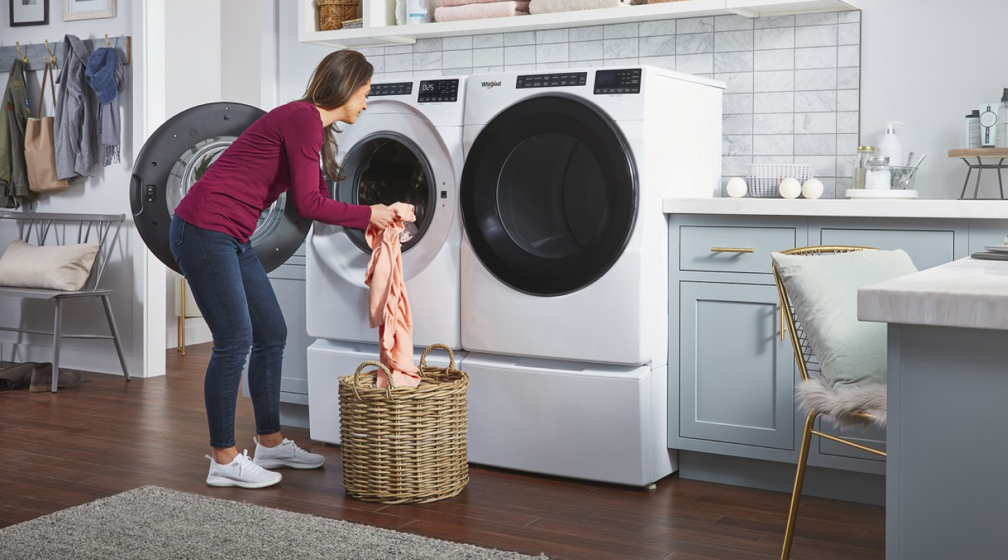 A woman putting laundry into a white Whirlpool® front load washer that sits next to a dryer in a modern laundry room. A woman putting laundry into a white Whirlpool® front load washer that sits next to a dryer in a modern laundry room.