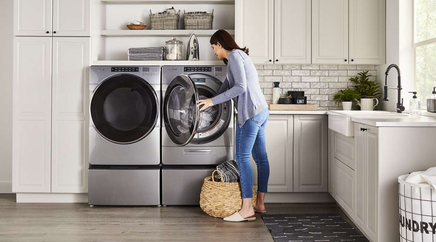 A woman putting laundry into a silver Whirlpool® front load dryer that sits next to a washer in a modern laundry room.