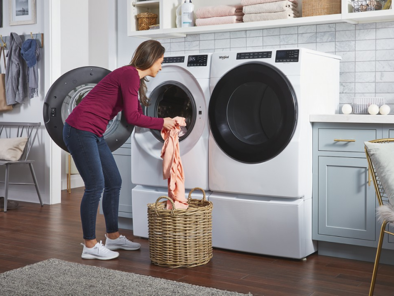 A woman putting laundry into a white Whirlpool® front load washer that sits next to a dryer in a modern laundry room. A woman putting laundry into a white Whirlpool® front load washer that sits next to a dryer in a modern laundry room.