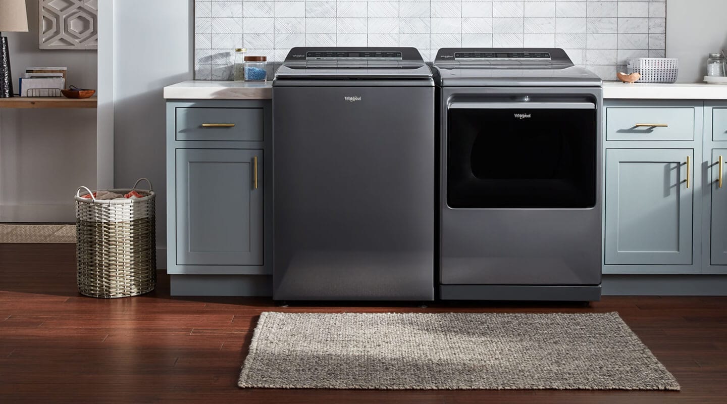A dark gray Whirlpool® Top Load Washer and Dryer in a laundry room with sky blue cabinets and white countertops