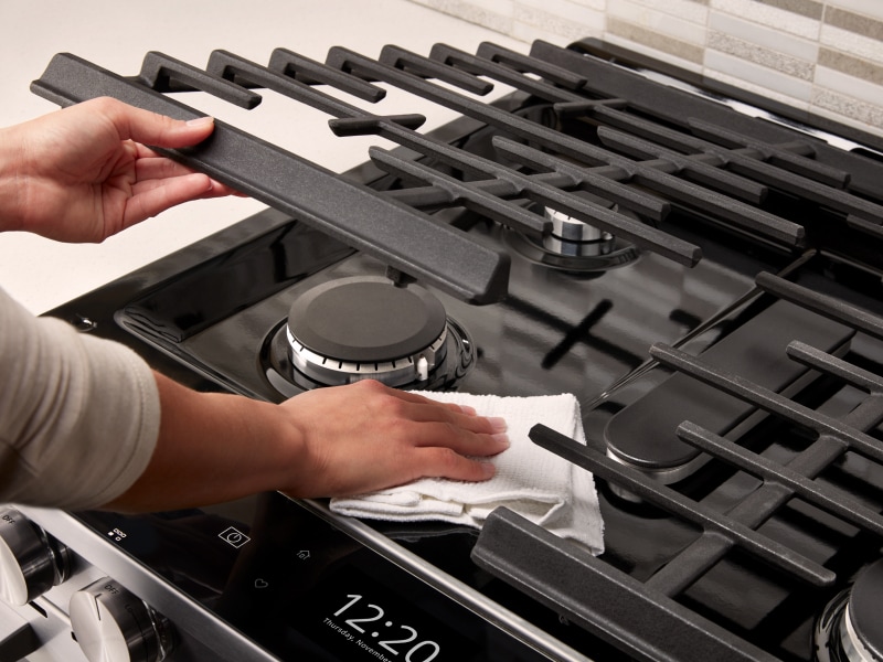 Person wiping underneath a gas cooktop grate Person wiping underneath a gas cooktop grate