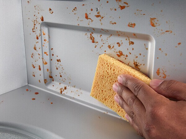 A person wiping food splattered on the interior of a microwave with a sponge