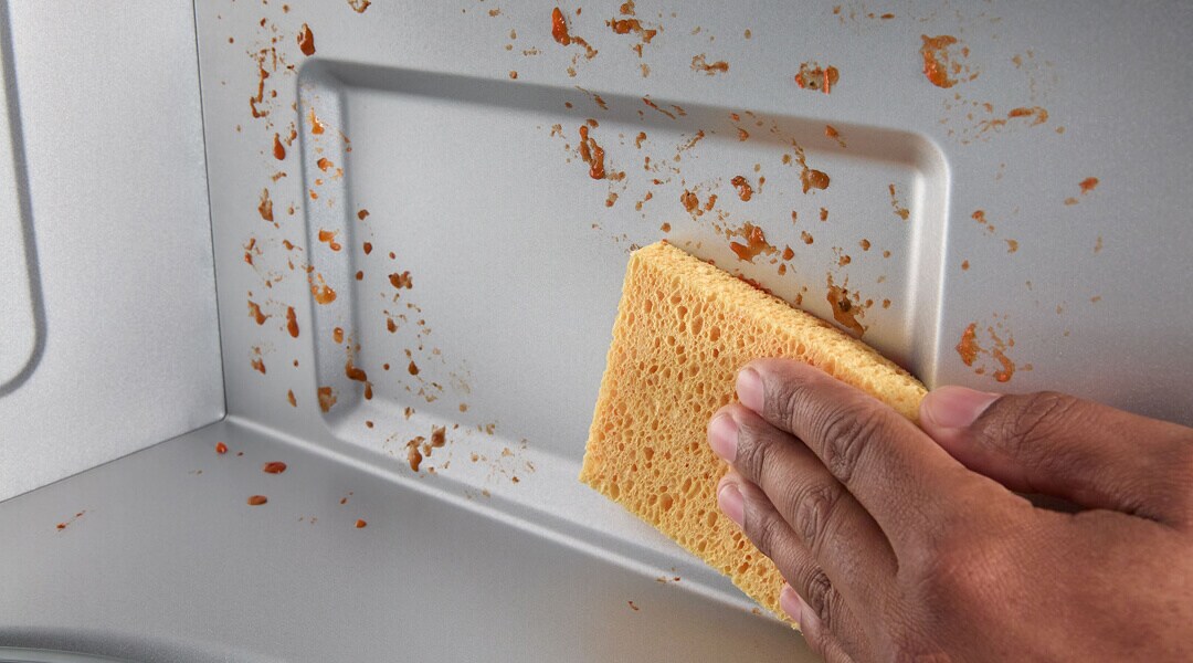 A person wiping food splattered on the interior of a microwave with a sponge