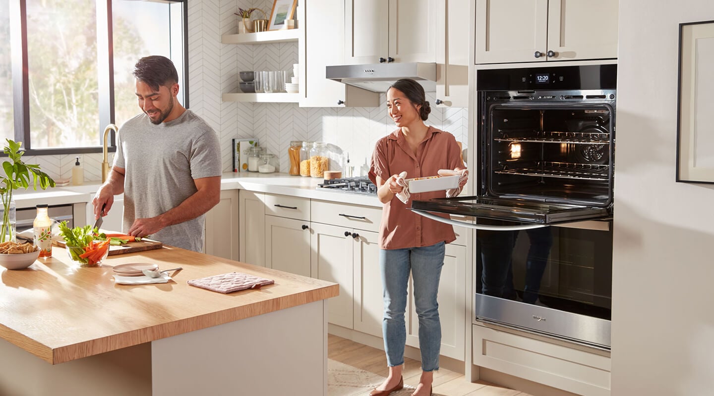 A man chopping vegetables at a kitchen island as a woman pulls food out of a wall oven