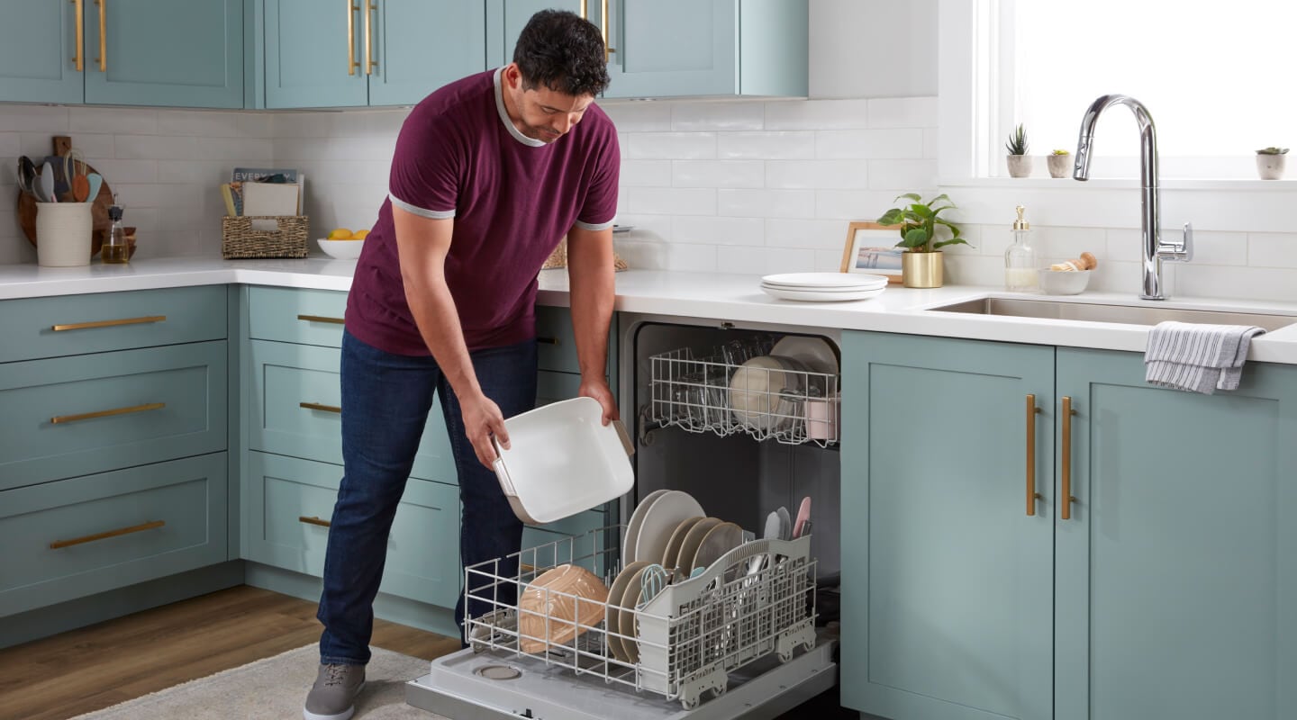 A person in a red shirt loading a built-in dishwasher in a mint green kitchen with white countertops and sink