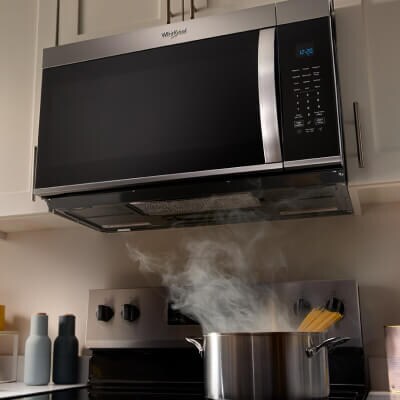 A pot releasing steam as it cooks on a stovetop below a microwave