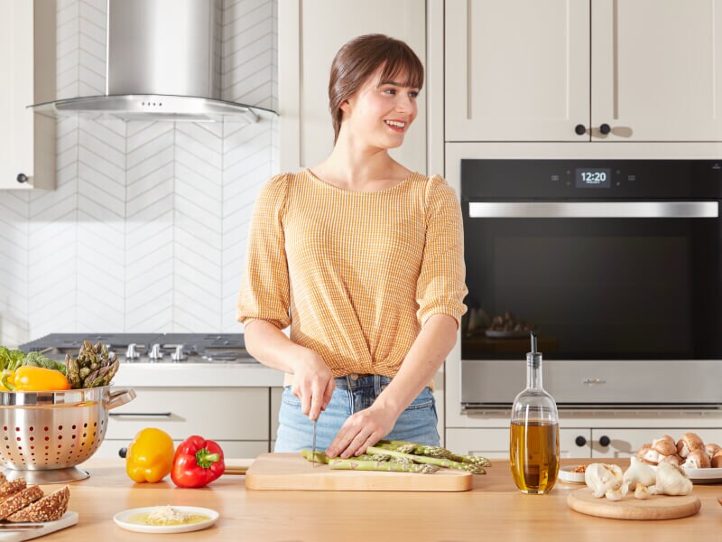 A person chopping asparagus on a wooden cutting board placed on a kitchen island with a cooktop in the background behind her A person chopping asparagus on a wooden cutting board placed on a kitchen island with a cooktop in the background behind her
