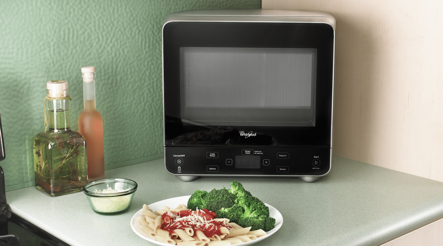 A Whirlpool® Countertop Microwave on a kitchen countertop next to a plate of pasta and broccoli. A Whirlpool® Countertop Microwave on a kitchen countertop next to a plate of pasta and broccoli.