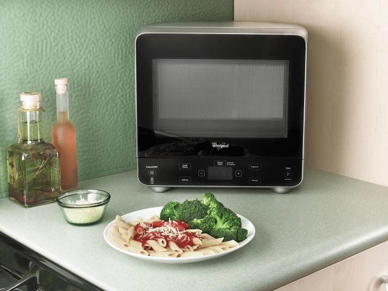 A Whirlpool® Countertop Microwave on a kitchen countertop next to a plate of pasta and broccoli. A Whirlpool® Countertop Microwave on a kitchen countertop next to a plate of pasta and broccoli.