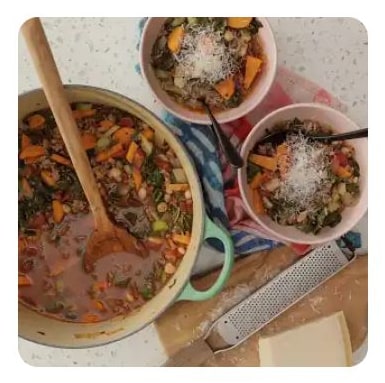 A pot and two bowls of vegetable beef soup with grated cheese
