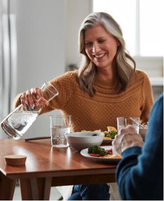 A woman fills up a clear glass with water from a full pitcher