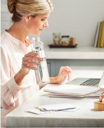 A woman drinks from a full reusable water bottle filled with clear water while working on the computer