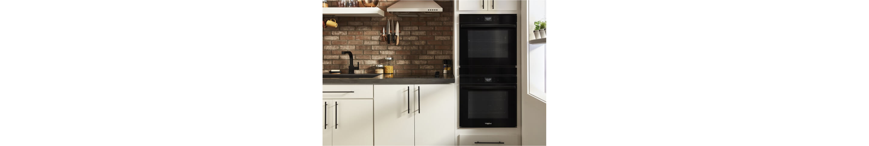 A Whirlpool® Small Space Double Wall Oven, Electric Cooktop and Wall Mount Range Hood in a kitchen with light cabinets