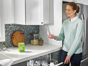 A person holding a glass next to an open dishwasher with a box of affresh® on the counter