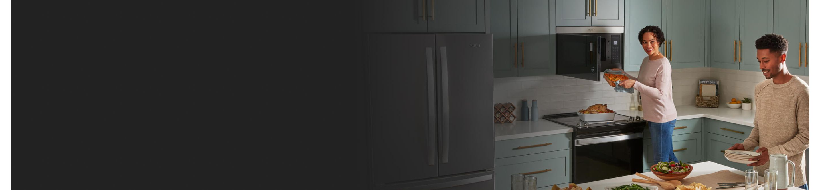 A couple making dinner in a sea green kitchen with one of them pulling a casserole dish out of a microwave